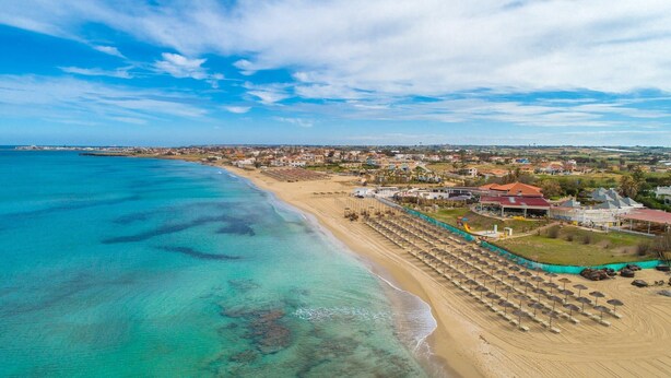 An aerial view of a coastal area with a beach, a row of umbrellas, and a building with a red roof.