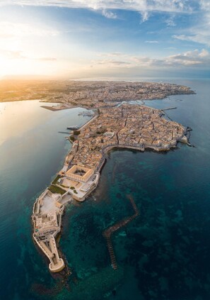 An aerial view of a coastal city with a prominent fortification along the shoreline, clear blue waters, and a distant cityscape.