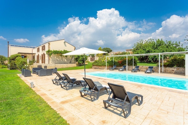 A pool area with lounge chairs and a pergola, surrounded by a well-maintained lawn and a large house in the background.