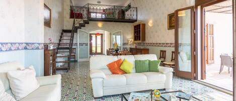 A spacious living room with a white sofa, colorful patterned floor, a glass coffee table, and a staircase leading to an upper level.