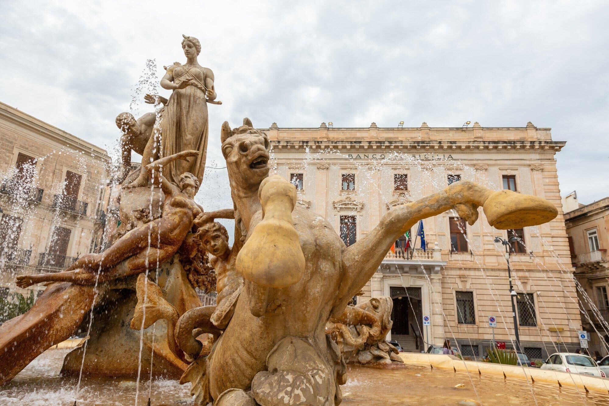A historic fountain with water jets and sculptures in front of a classical building with columns and a balcony.