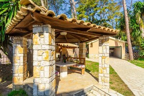 Shaded outdoor seating area beneath a pergola with a wooden roof and stone pillars.