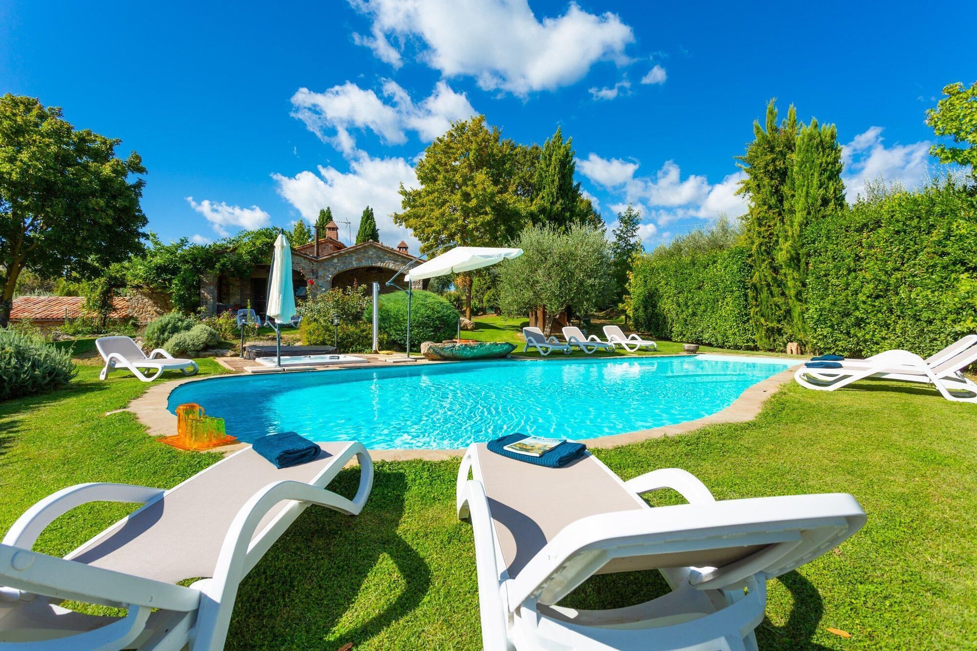 A swimming pool with lounge chairs and umbrellas, surrounded by greenery and a stone building.