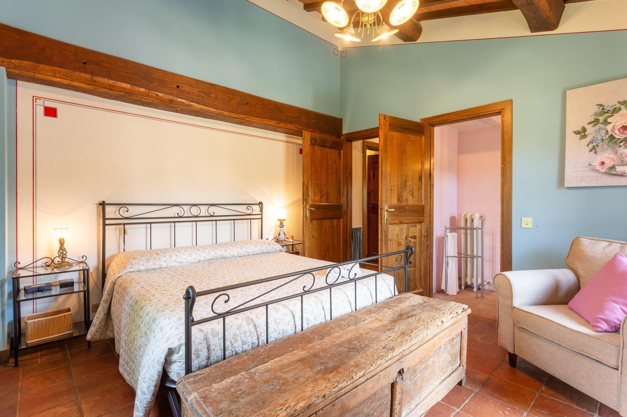 Un dormitorio con estructura de cama de madera, una mesita de noche con lámpara, una silla con cojín y un baúl de madera.