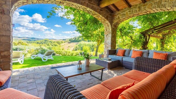 Un espacio de estar al aire libre cubierto con arcos de piedra, vistas al campo y una mesa con un jarrón de flores.