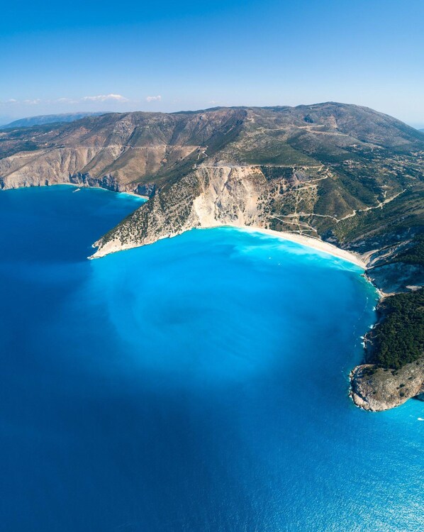 An aerial view of a coastal area with a clear blue sea, a sandy beach, and rugged terrain.