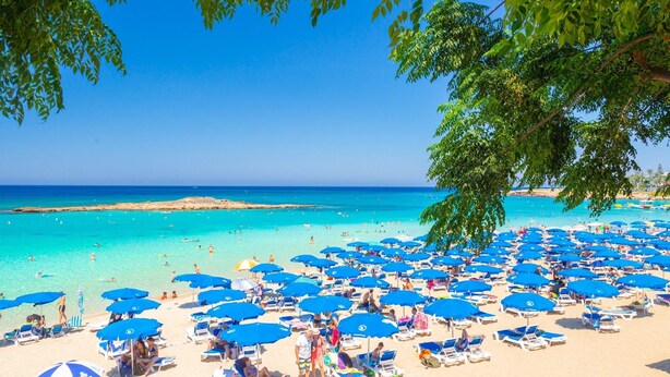 Une plage parsemée de nombreux parasols bleus et de chaises longues, un ciel bleu dégagé et une mer calme.