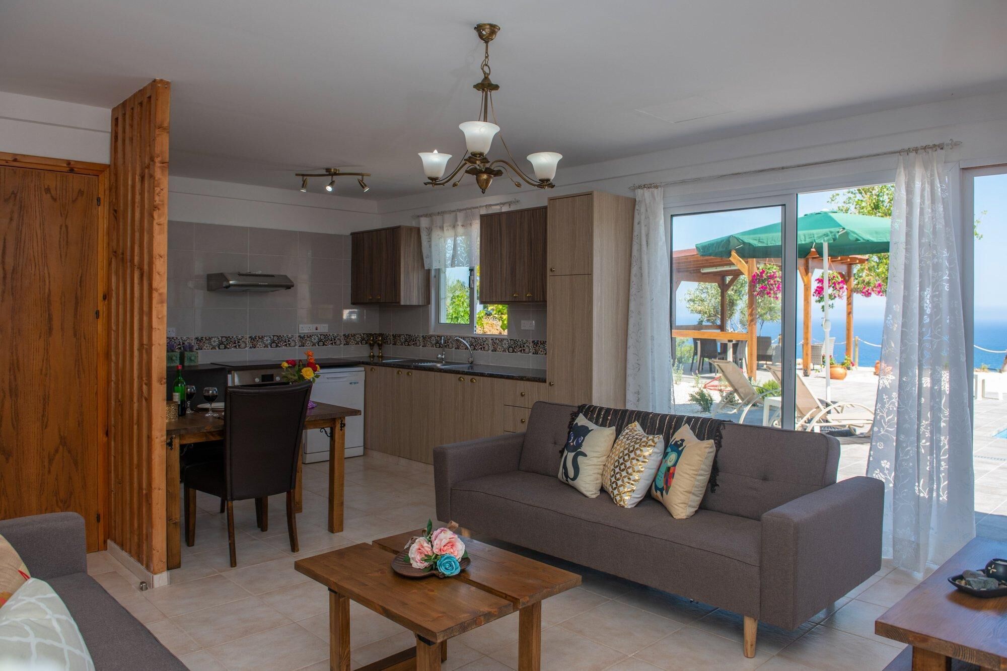 Un appartement moderne comprenant une cuisine, un coin repas et un salon. Il dispose d’une grande fenêtre offrant une vue sur la mer, d’une table à manger en bois, d’un canapé et d’une petite table basse.