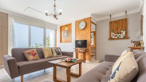 Un salon moderne avec un canapé, une table basse et un téléviseur. Un panneau mural en bois est agrémenté d’une horloge et d’une guirlande décorative.