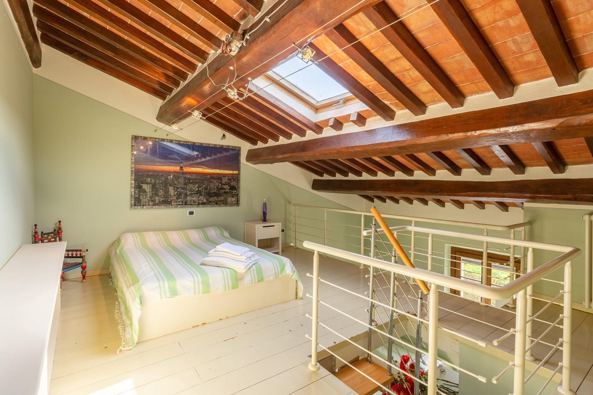 Un dormitorio con una cama, un tragaluz, un cuadro de la ciudad y una escalera.
