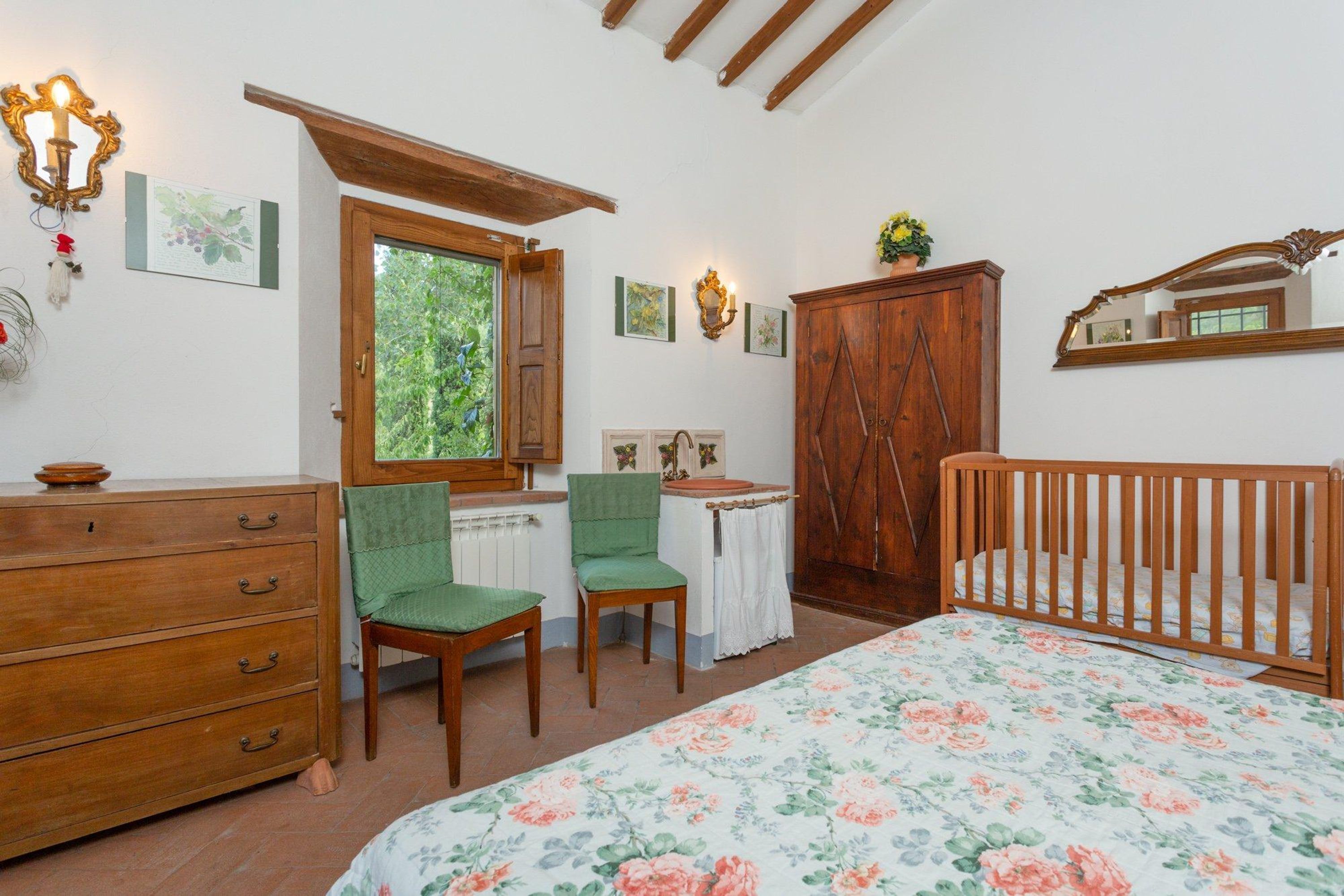 Een slaapkamer met een houten bed, een kapstok, twee stoelen, een bureau en een raam met uitzicht op groen.