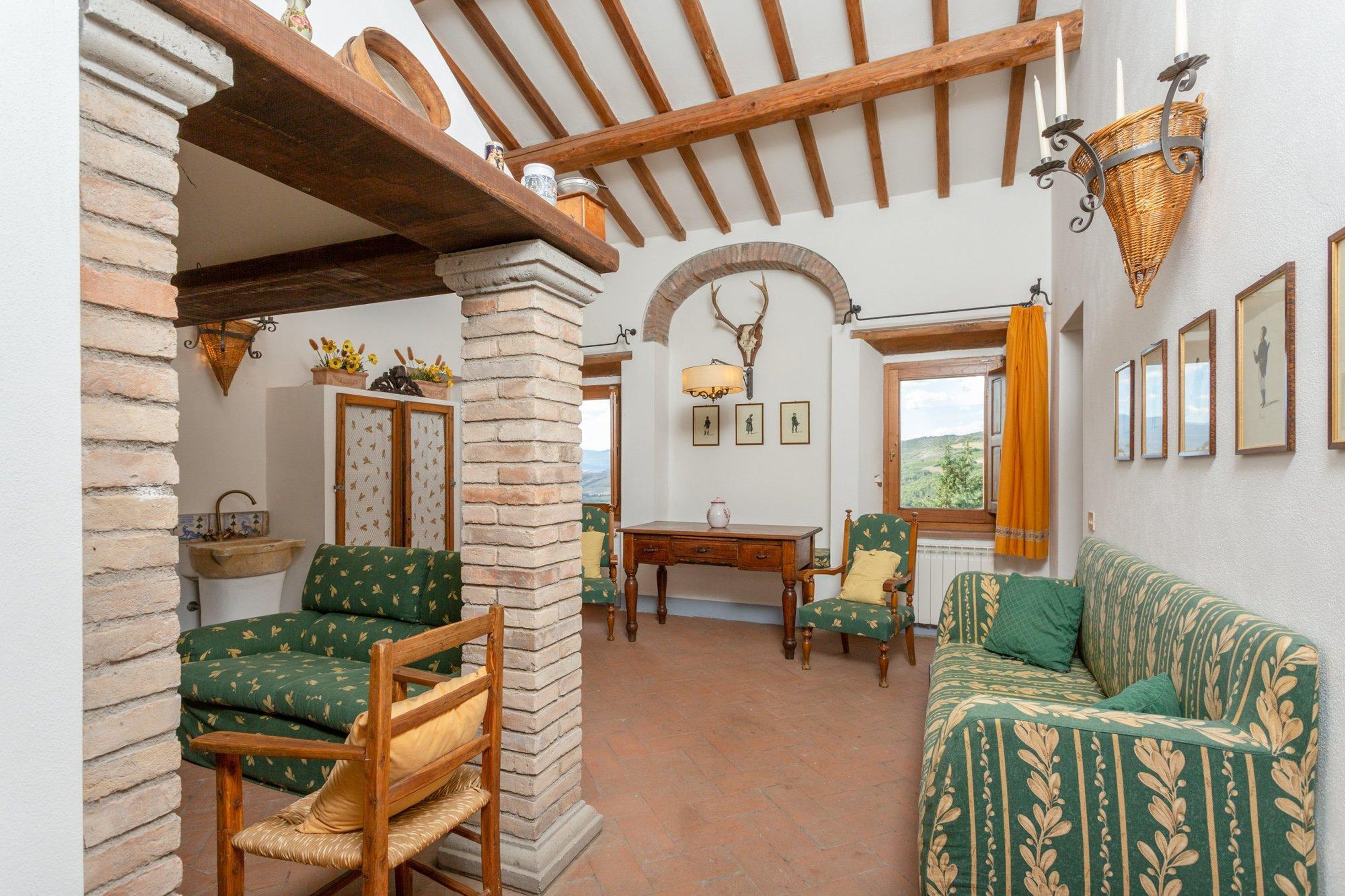 A rustic interior with exposed wooden beams, a stone pillar, a green patterned sofa, a wooden chair, a table, and a wall-mounted deer head.