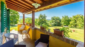 Une terrasse couverte, aménagée avec des meubles en osier, des plantes en pot et offrant une vue sur un paysage verdoyant.