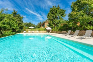 En klarblå pool med solstolar och parasoller, omgiven av frodig grönska och en stenbyggnad i bakgrunden.