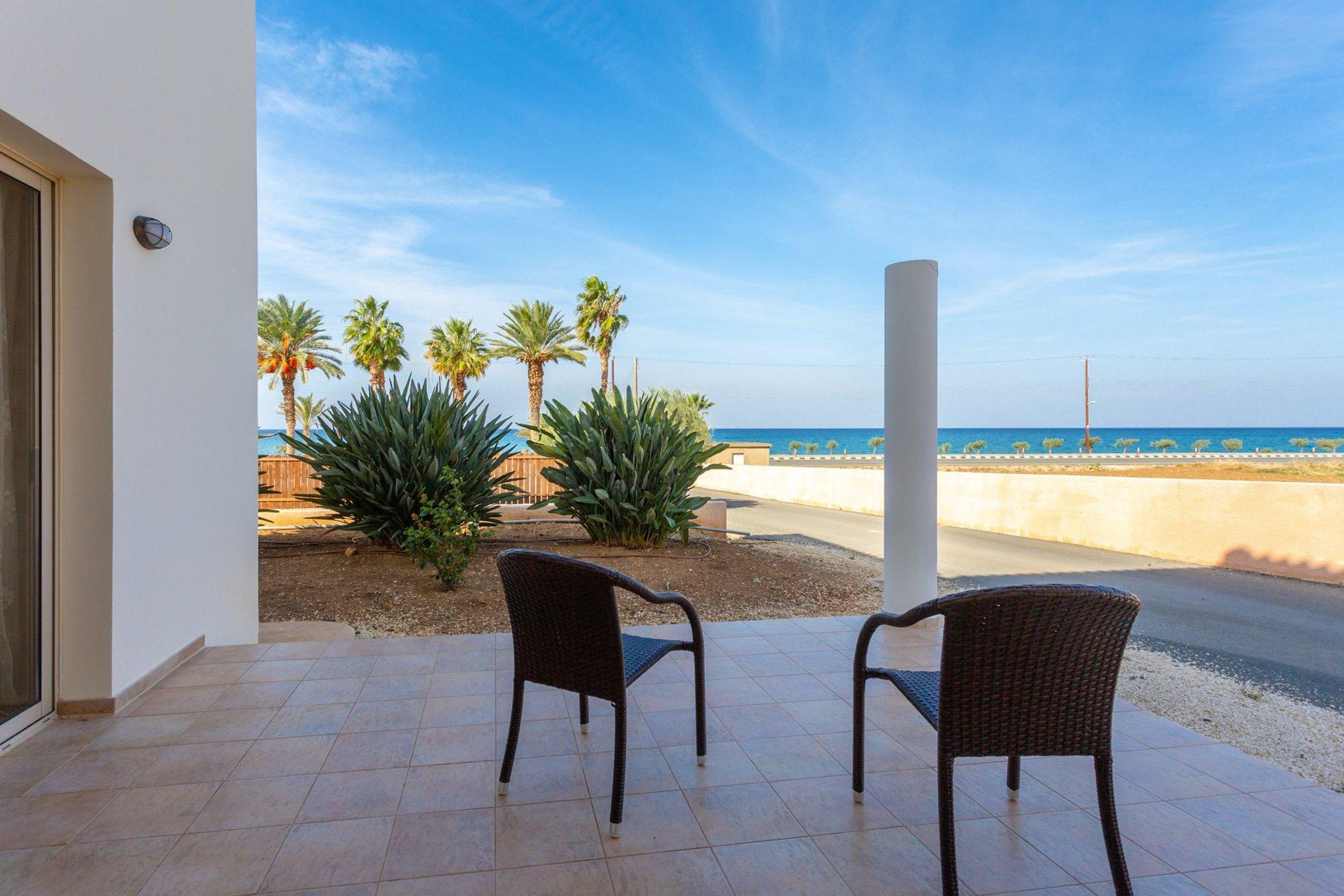 Un patio con dos sillas de mimbre mirando al mar, palmeras y un pilar blanco.