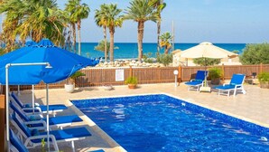A swimming pool with blue water, surrounded by lounge chairs and umbrellas, palm trees, and a view of the sea. - Villa Seashore One Large Private Pool Walk to Beach Sea Views A C Wifi - 3150 (Argaka)