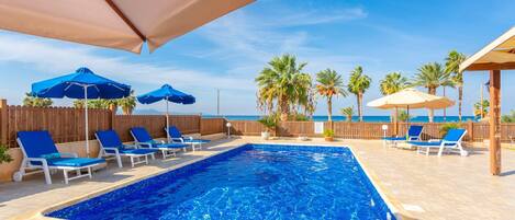 A rectangular swimming pool with blue water, surrounded by lounge chairs and umbrellas, with palm trees and a wooden fence in the background.