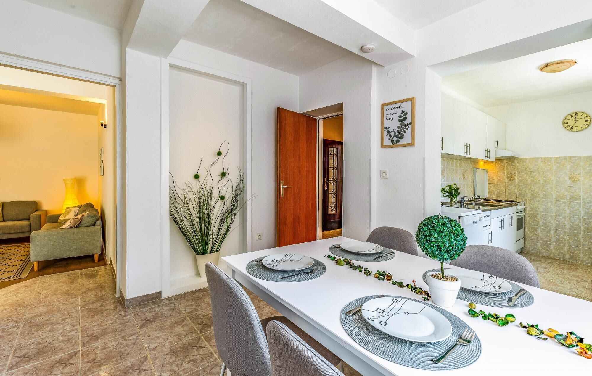 A dining area with a white table set for four, a kitchen visible through an open door, and a living area with a sofa and lamp.