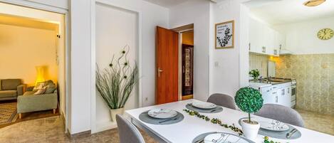 A dining area with a white table set for four, a kitchen visible through an open door, and a living area with a sofa and lamp.