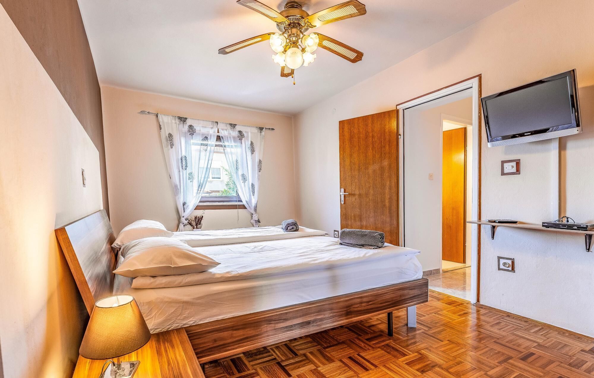 A bedroom with a wooden bed, a ceiling fan, a TV mounted on the wall, and a window with curtains.