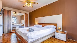 Habitación de hotel con piso de madera, una cama con sábanas blancas, una mesita de noche con lámpara y un ventilador de techo.