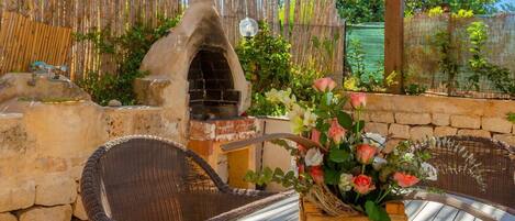 An outdoor patio with a wooden pergola, a stone wall, a brick oven, wicker chairs, a table with a tray of food, and a flower arrangement.