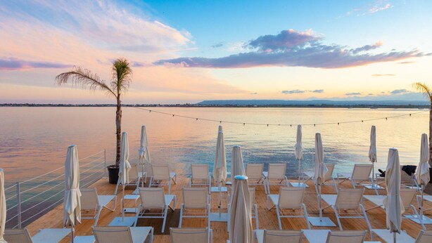 A deck with rows of white lounge chairs and umbrellas, overlooking a calm body of water with a sunset sky.