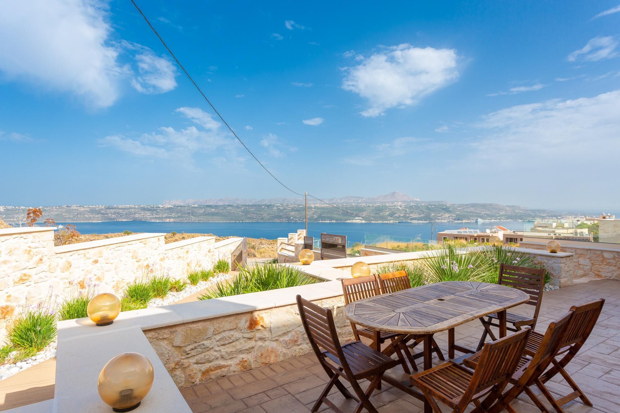 Eine Terrasse mit rundem Tisch und Stühlen, mit Blick auf ein malerisches Gewässer und ferne Hügel.