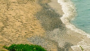 Ein Strand mit Kieselsteinen und Sand, ein großer Felsen und eine Person am Wasser.