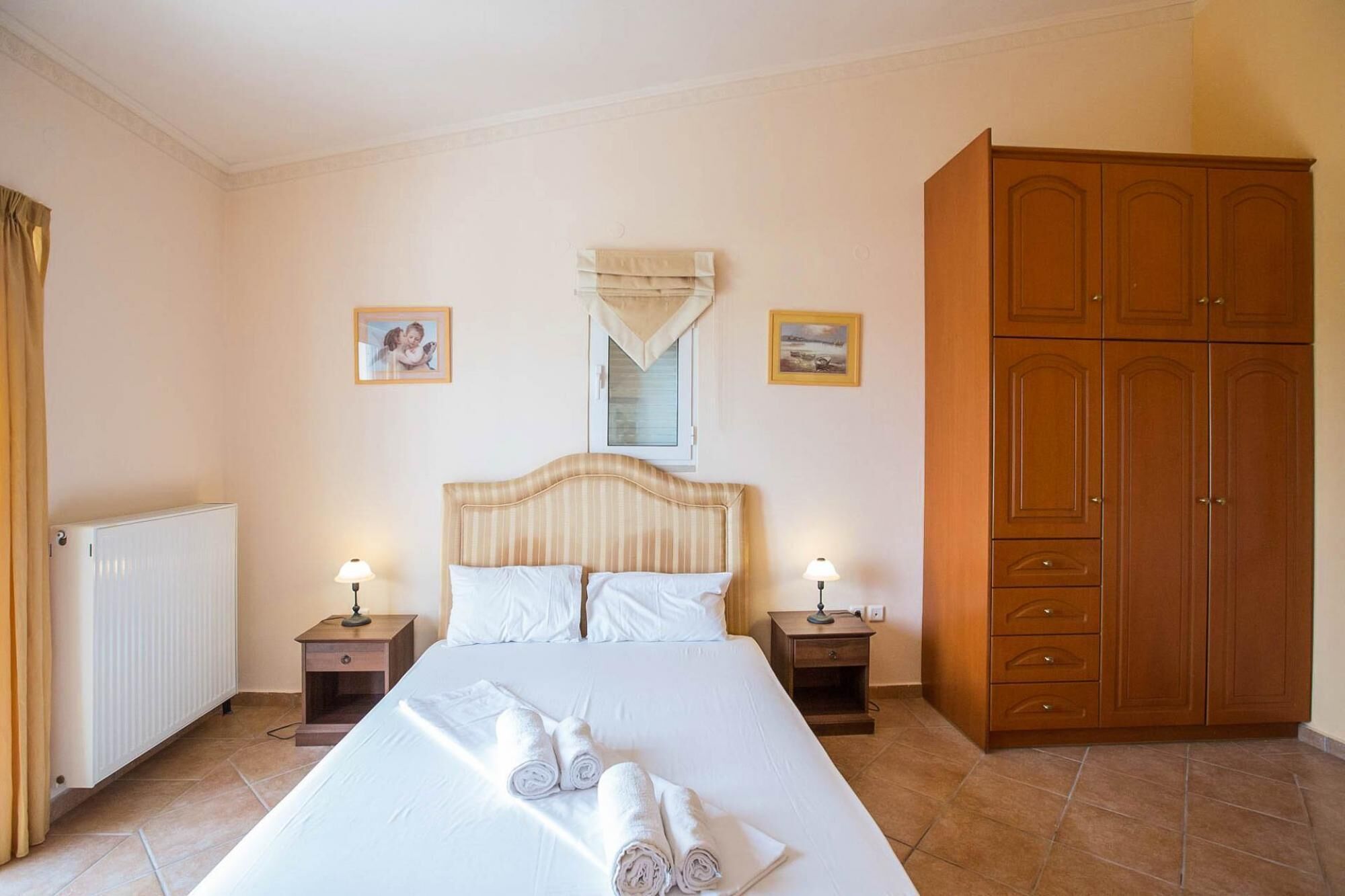 A hotel room with a large bed, two bedside tables, a wooden wardrobe, and two framed pictures on the wall.