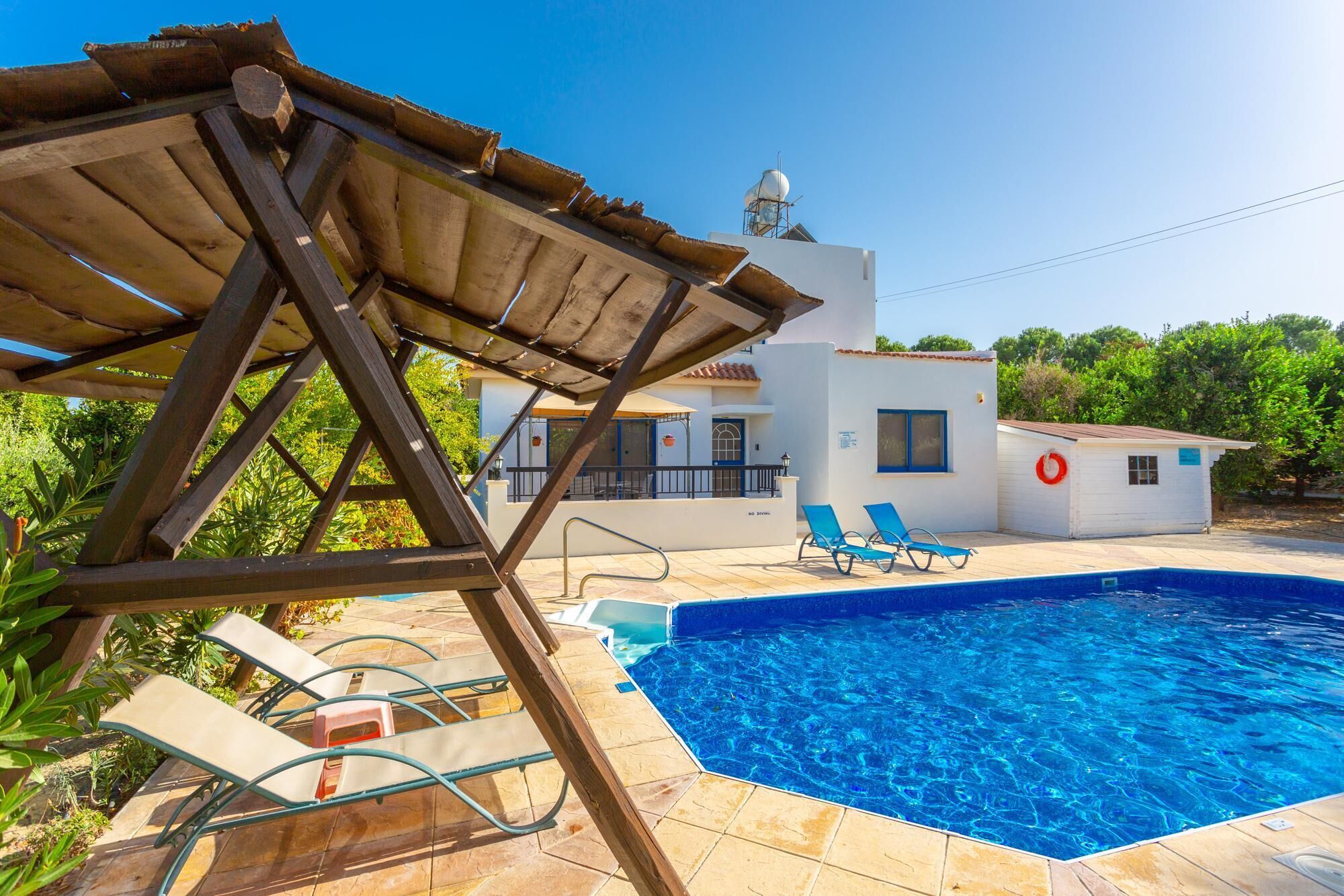 A swimming pool with lounge chairs, a wooden pergola, and a white building with a blue door.