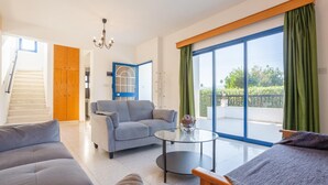 A spacious living room with a grey sofa, a blue armchair, and a glass coffee table. There is a wooden door with a blue frame, a staircase, and a large window with green curtains. - Villa Charoula Exi Large Private Pool A C Wifi - 3275 (Polis)