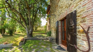 Un bâtiment en pierre doté d’une porte et de volets en bois, entouré d’un chemin de pierre et d’une végétation luxuriante.