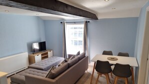 Ein kompakter Wohnraum mit einer Couch, einem Fernseher, einem Esstisch mit Stühlen und einem Fenster mit Vorhängen.