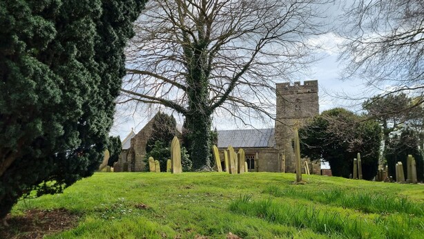 Ein Friedhof mit Grabsteinen, einem großen Baum und einem Steinhaus im Hintergrund.