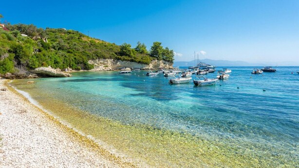 Una playa con agua turquesa cristalina, orilla pedregosa y barcos fondeados cerca.