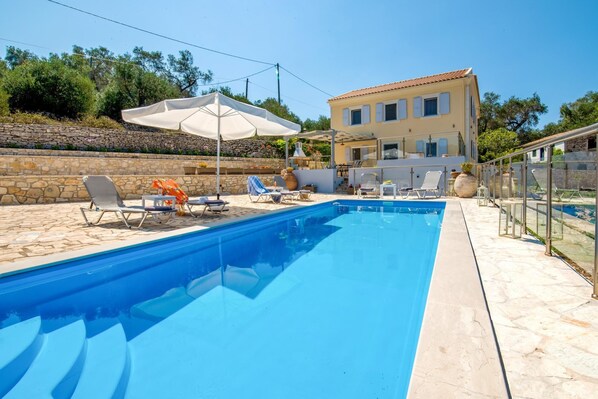 A swimming pool with lounge chairs and umbrellas, a stone wall, and a building in the background.