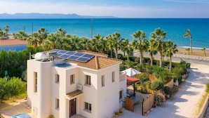 A modern two-story building with a rooftop solar panel array, surrounded by palm trees and a beach view.