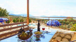 A table with food and drinks, a bottle, and a view of mountains. - Villa Yeraki Large Private Pool Sea Views A C Wifi - 981 (Kefalonia)