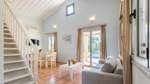 A spacious living area with a white staircase, a dining table set with chairs, a sofa, and a large window with curtains. - Antigoni Beach House Large Private Pool Walk to Beach Sea Views A C Wifi Car Not Required - 1012 (Kefalonia)