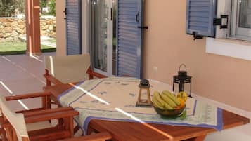 Uma mesa de madeira com toalha florida, uma tigela de frutas e duas lanternas numa varanda com persianas azuis.