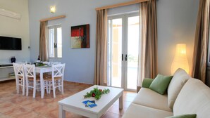 A living room with a white sofa, a coffee table with a plant, a dining area with white chairs, and a painting on the wall. - Nafsika Beach House Large Private Pool Walk to Beach Sea Views A C Wifi Car Not Required - 1880 (Kefalonia)