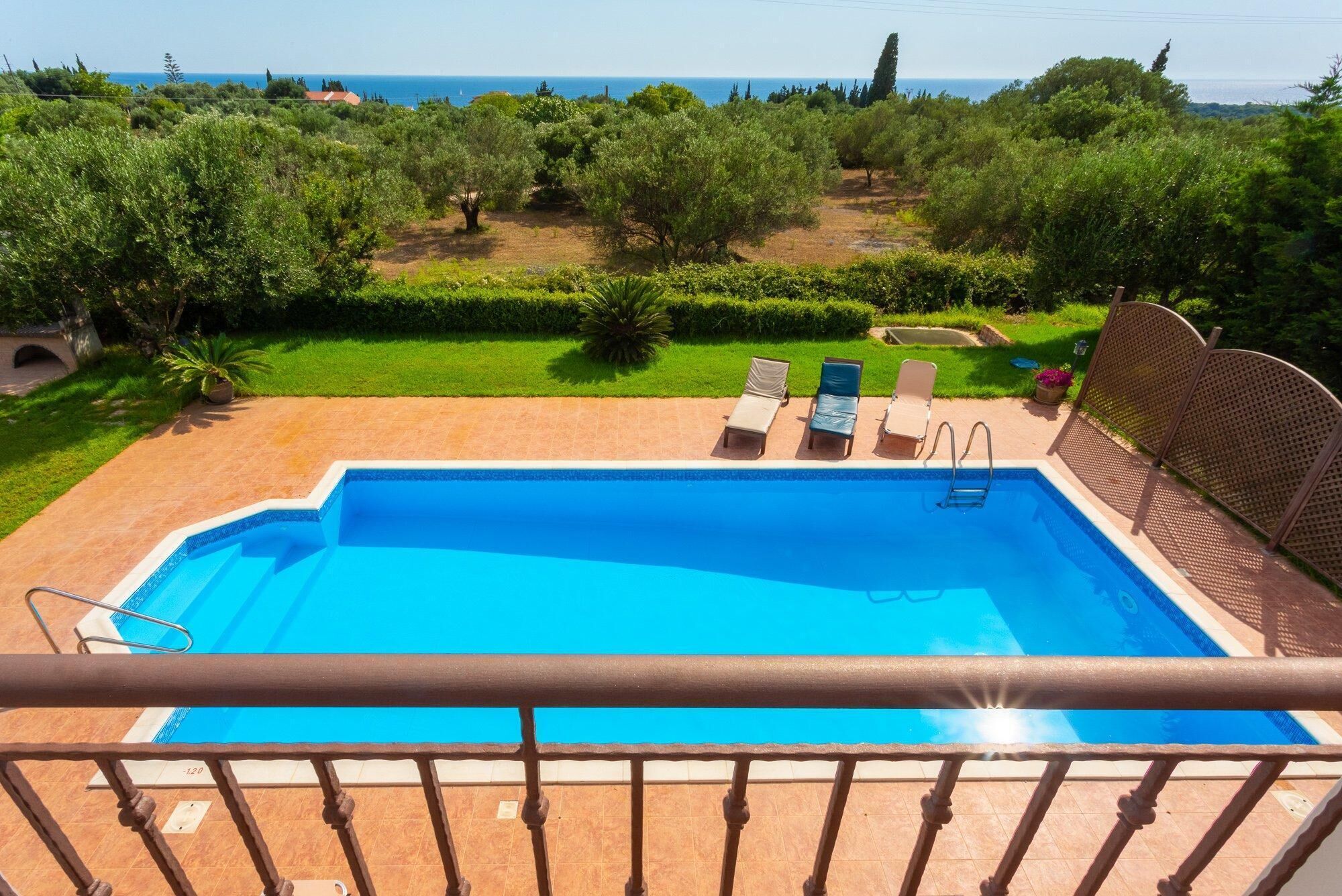 A swimming pool with a view of the sea, surrounded by greenery and a fenced area with lounge chairs.