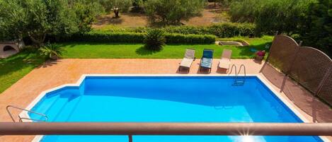 A swimming pool with a view of the sea, surrounded by greenery and a fenced area with lounge chairs.