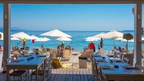 Ein Essbereich im Freien mit Tischen und Stühlen, mit Blick auf einen Strand mit Sonnenschirmen und Personen.