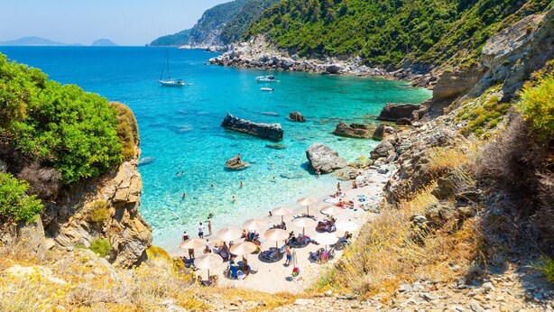 A coastal beach with clear turquoise waters, rocky cliffs, and lush greenery.