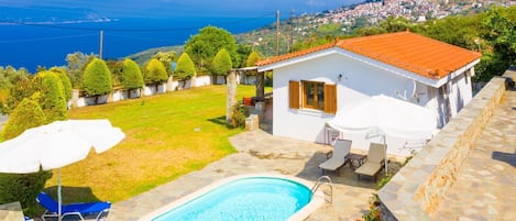 A white villa with a swimming pool, outdoor seating, and a view of the sea and town.