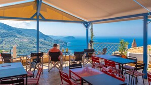 An outdoor terrace with tables and chairs, overlooking a coastal landscape with hills and the sea.