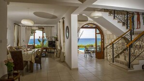 A spacious dining area with a view of the sea, a staircase with ornate railings, and a cozy seating area with a TV. - Villa Minoas Large Private Pool Walk to Beach Sea Views A C Wifi Eco-friendly - 2565 (Argaka)