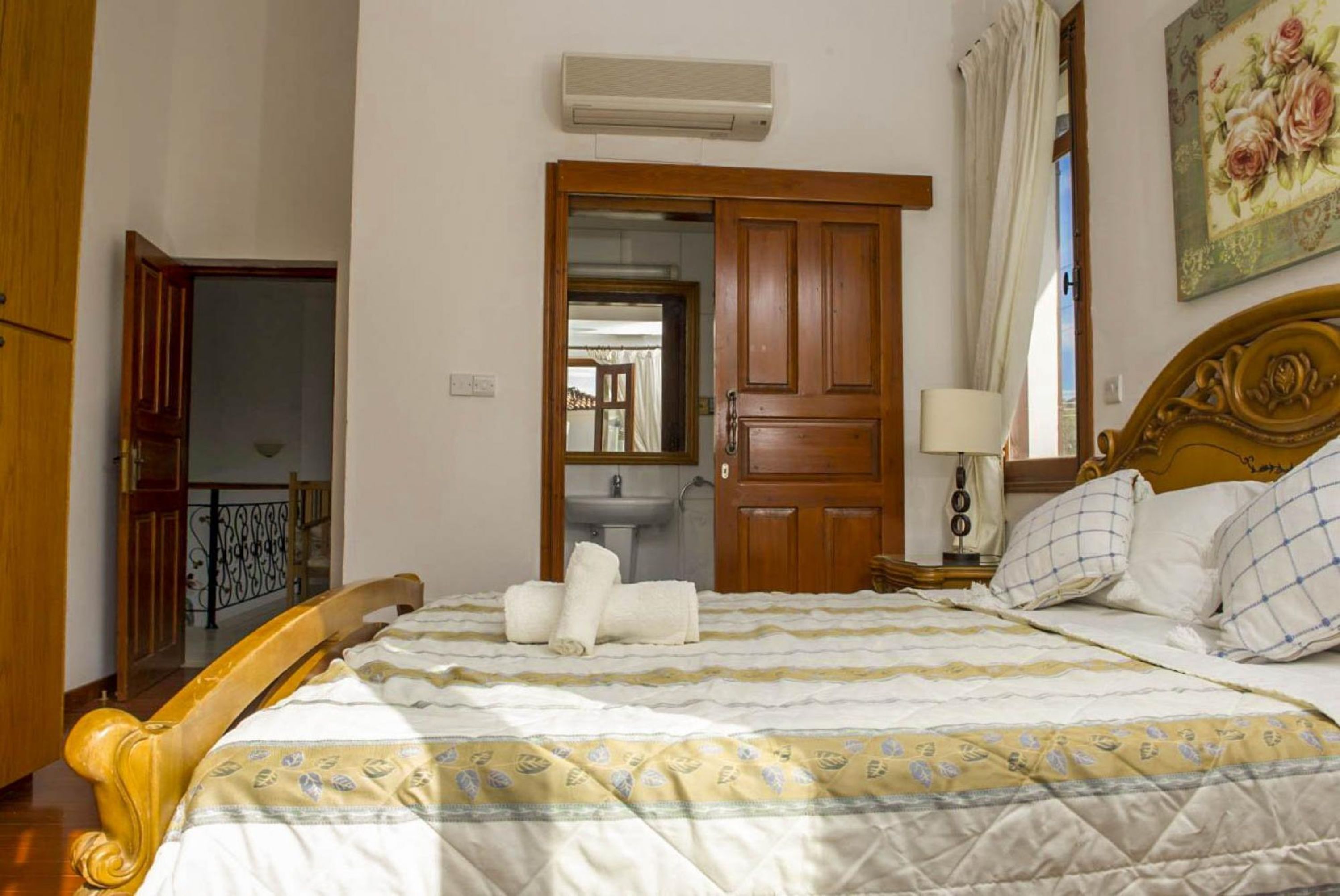 Une chambre à coucher avec un lit en bois, une table de chevet avec une lampe, une salle de bain visible à travers une porte ouverte, et un tableau accroché au mur.